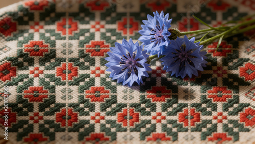 Three blue cornflowers on a cream linen runner with red and green ethnographic pattern, Latvian Midsummer still life.