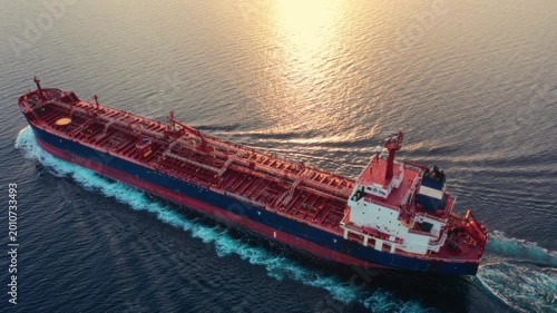 Large crude oil tanker underway on open sea at golden hour, aerial view showing full vessel length with pipeline deck and bow wave. Aerial view