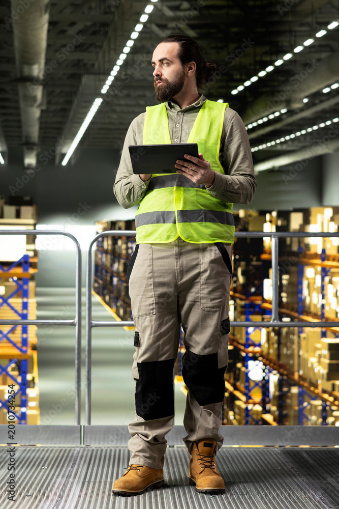 Obraz premium Adult man working on tablet on warehouse platform in distribution center, checking shipment and transportation details. Managing cargo and logistics for order fulfillment and control.