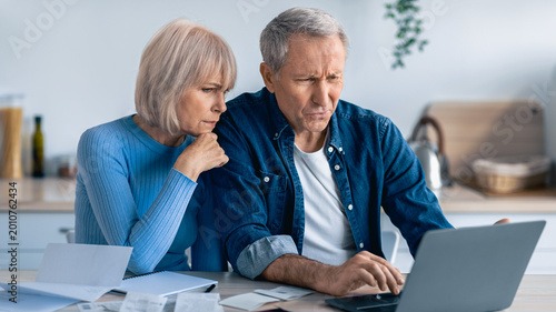 Slika na platnu Two older adults are seated at a table filled with papers, looking at a laptop