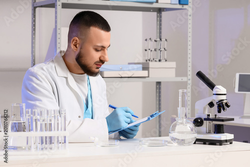 Male scientist with glassware of water samples writing in clipboard at labora...