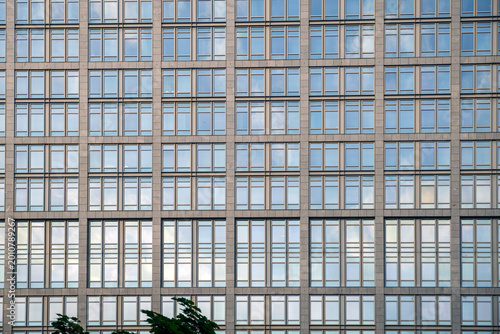 Low angle view of a massive glass and steel skyscraper facade reflecting the sky with tree branches in the foreground.