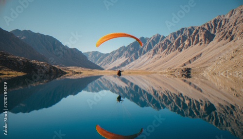 Foto A paraglider gliding over a calm mountain lake with a perfectly symmetrical reflection in the water, surrounded by rugged, rocky mountains under a clear blue sky