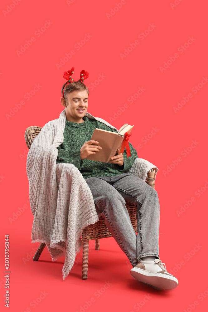 Fototapeta premium Young man reading book while sitting in armchair on red background. Christmas celebration