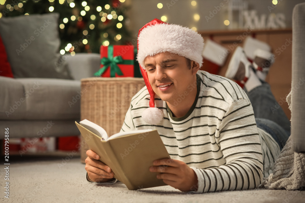 Fototapeta premium Young man in Santa hat reading book at home on Christmas eve