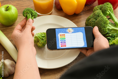 Young woman using meal plan app on mobile phone and holding fresh broccoli on...