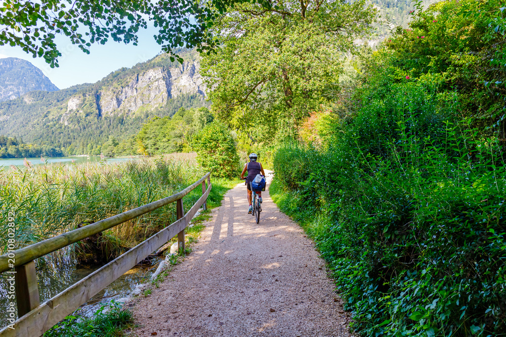 Fototapeta premium Biker rides along a pathway by the lake surrounded by trees and mountains during a sunny day in nature