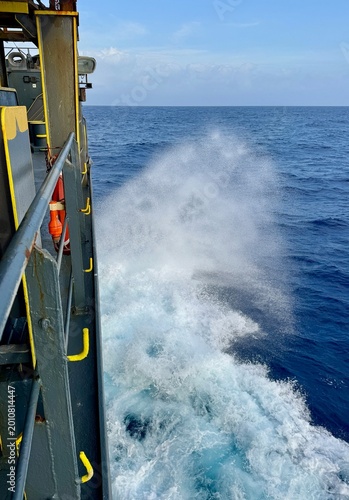 Side view of a vessel showing metal structure and railing with powerful ocean spray hitting the side, surrounded by deep blue water, representing industrial shipping and life at sea