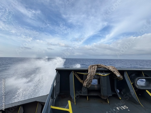 View from the bow of a moving ship cutting through ocean waves, with strong water spray on both sides, dramatic cloudy sky, and visible deck structure emphasizing motion, power