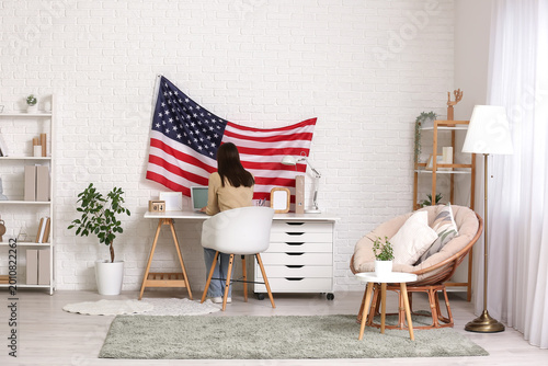 Young woman using laptop at workspace with USA flag on wall