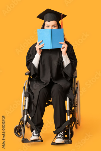 Beautiful female graduate student in wheelchair with graduation cap reading b...