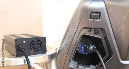 Modern charge EV vehicle electric battery charger plugged into motorcycle port. Clean metallic design highlights advanced technology. Charging port open, cable connected with blue connector.