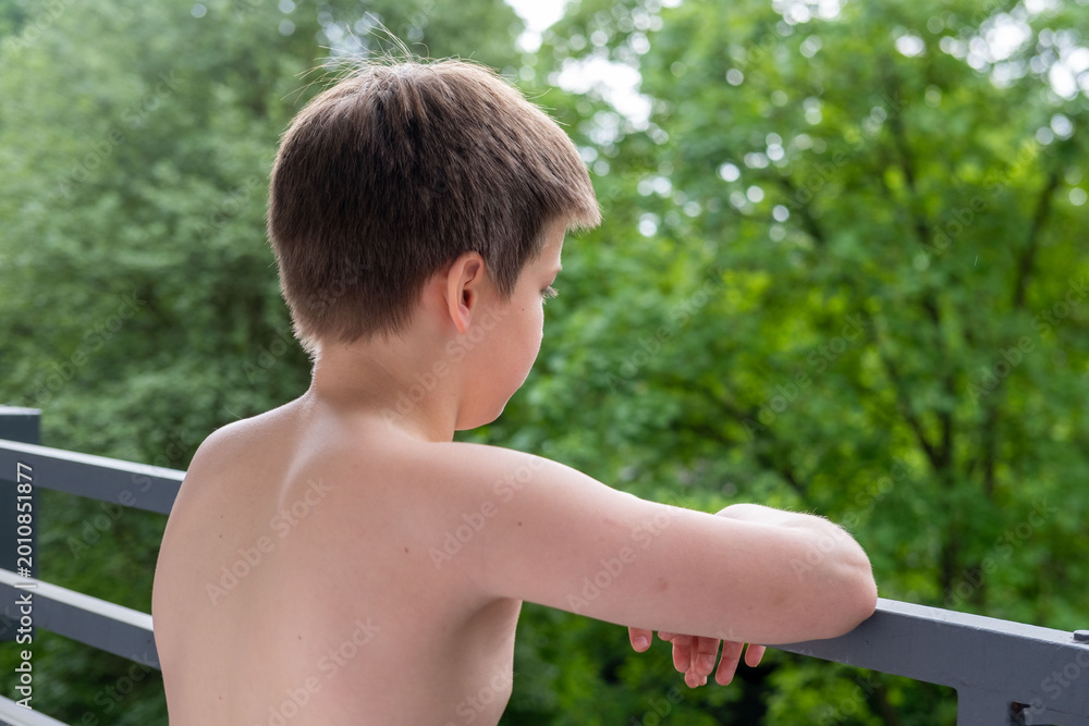 Naklejka premium Young Shirtless boy standing on terrace, balcony, enjoying fresh air outdoors, summer day, peaceful Back view, green forest background, childhood moment, nature connection