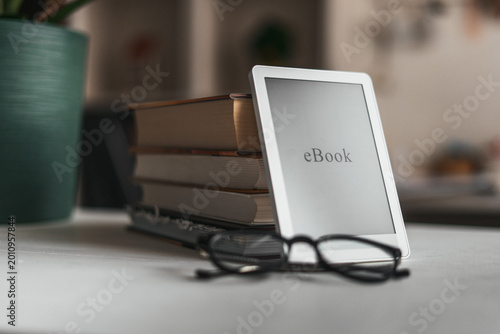 Ebook reader and glasses on desk with books and plant in soft home office setting. Cozy workspace, remote work lifestyle and calm productivity concept with minimalistic interior design.