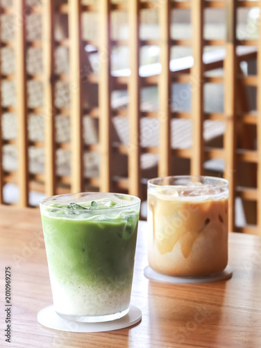 Two refreshing iced beverages on a wooden table in a cozy cafe setting: a layered iced matcha latte with creamy milk and a smooth iced coffee latte.
