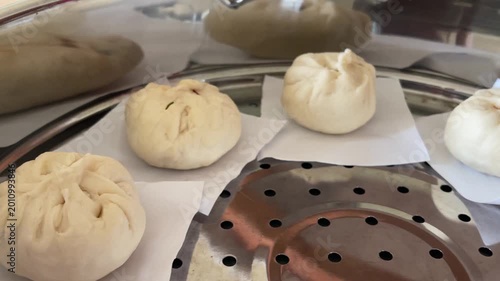 A horizontal panning shot showcases an array of freshly pleat-wrapped raw baozi buns neatly organized on parchment paper inside a steamer tray, ready for steaming.
