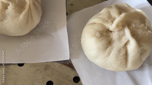 A panning shot reveals several freshly pleat-wrapped raw baozi buns neatly arranged on steamer paper inside a traditional steamer, ready for cooking.