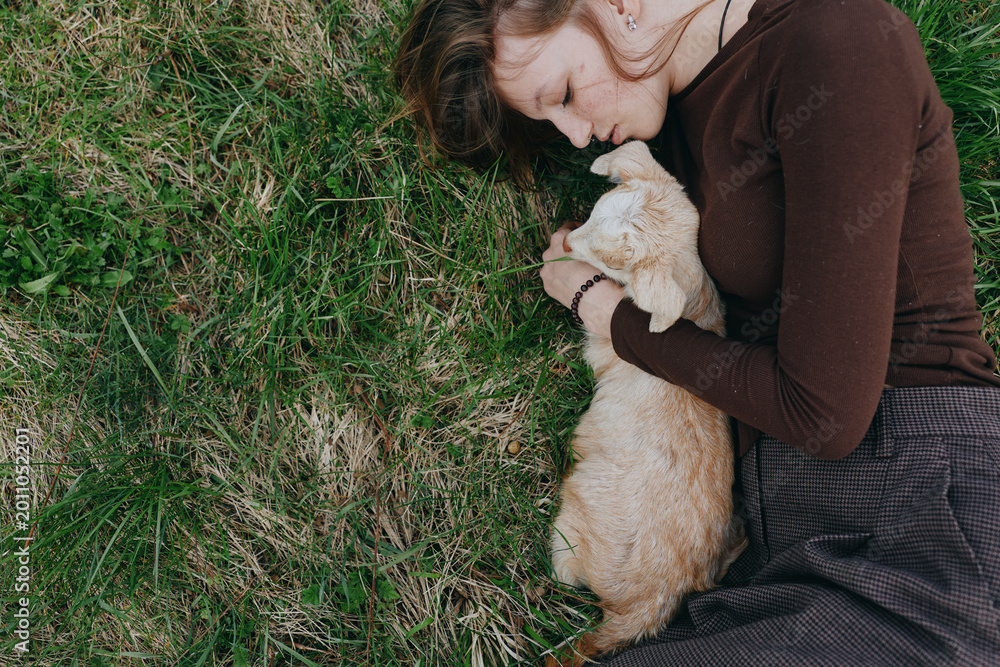 Naklejka premium Carefree woman on grass gently cuddling a young goat kid, capturing a tranquil countryside moment. Natural light highlights affection, nature, and simple rural living.