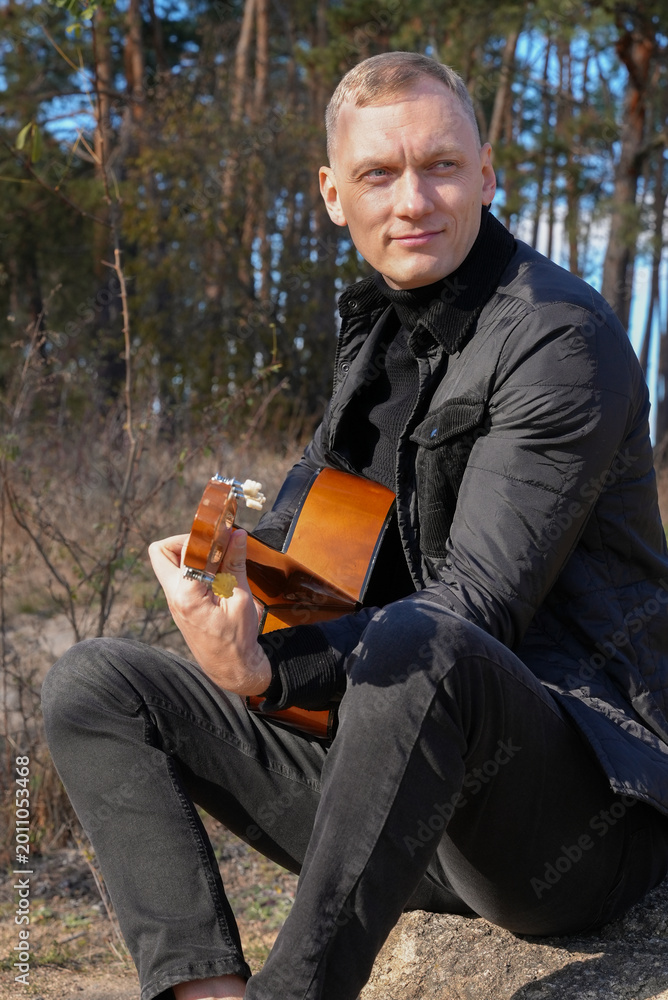 Obraz premium Beautiful blonde man playing acoustic guitar and singing relaxing enjoying outdoors on sunny autumn day. Artist learning and practicing playing musical instrument. Fall leaves autumnal weather 