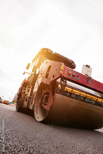 Workers are using a steamroller to pave asphalt on a busy urban roadway. The machinery is positioned on the surface, and the sky is partly cloudy, indicating day work being done.