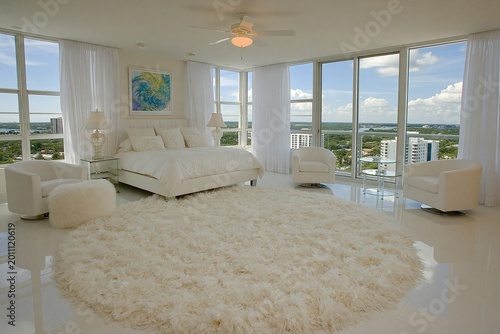 Bedroom offers a bright and airy atmosphere featuring minimalist white furniture, a large circular shaggy rug, and floor-to-ceiling windows that provide an expansive view of the surrounding city.