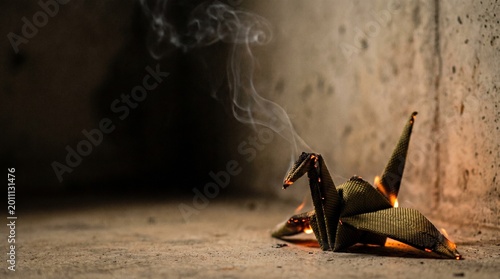 Burning origami crane folded from woven kevlar fabric smoldering on a dark concrete floor with smoke and copy space, concept of fragile peace and conflict
