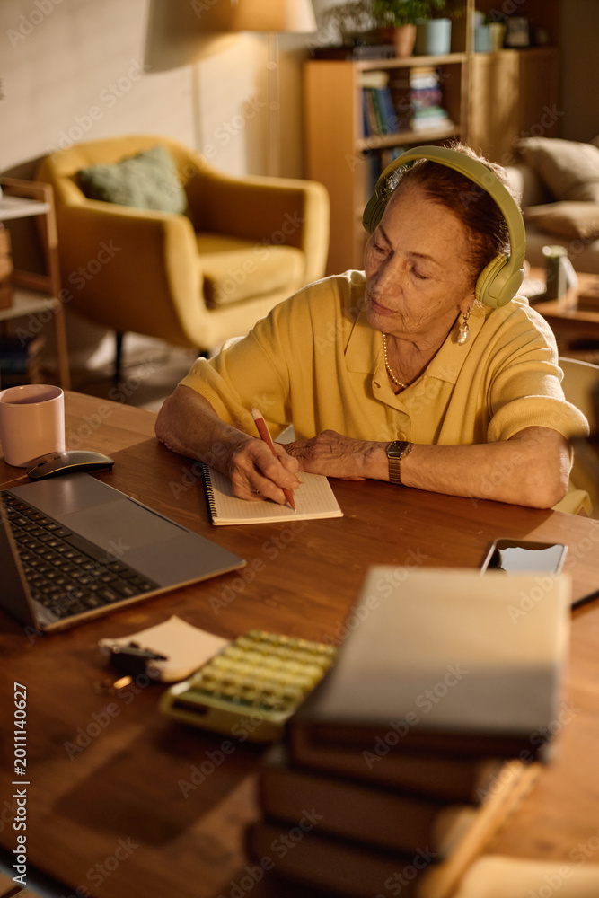 Obraz premium Vertical shot of senior Caucasian woman wearing headphones, writing notes beside laptop and calculator at home. Supporting online learning, remote training, financial planning content