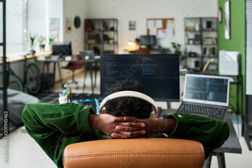 Tableau sur toile Young African American man reclining at coding workstation, wearing headphones and resting after programming session