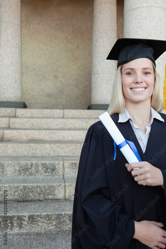Fototapeta premium Woman wearing graduation gown and cap holding diploma with blue ribbon on right steps, copy space