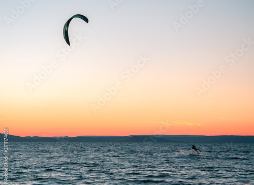 Kitesurfer rides ocean waves with a vibrant kite under a beautiful gradient sunset sky and distant mountains