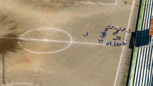 Konstfotografi High-angle shot of a group of people in blue uniforms gathered on a dirt sports field with white markings, including a center circle, next to concrete bleachers under bright, direct sunlight