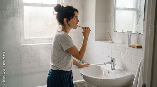 Person in bathroom brushing teeth at plain white sink in soft steam-filled morning light with white tiles