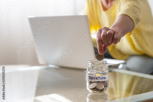 Person dropping a coin into a glass jar labeled donation while working on a laptop, symbolizing online fundraising, charitable giving, and financial support from home