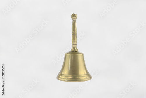 Golden bell stands alone against plain white background. Its handle curves upward, topped with a rounded knob. Bell surface gleams under soft even lighting. Vintage brass hand bell traditional  design