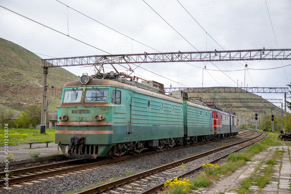 Fototapeta premium Electric freight locomotive, a vl10 model, waiting on tracks at a provincial railway station in armenia, with an overhead line system and hills in the background on an overcast day