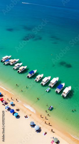 Aerial view of a tropical sandy beach with numerous white motorboats anchored in clear turquoise water and colorful umbrellas
