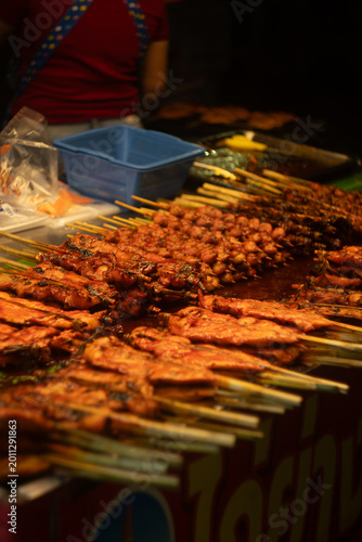 Vibrant close-up of a bustling Thai street food stall at night, featuring rows of freshly grilled meat skewers glistening under warm lighting. The juicy, caramelized skewers (chicken and pork).