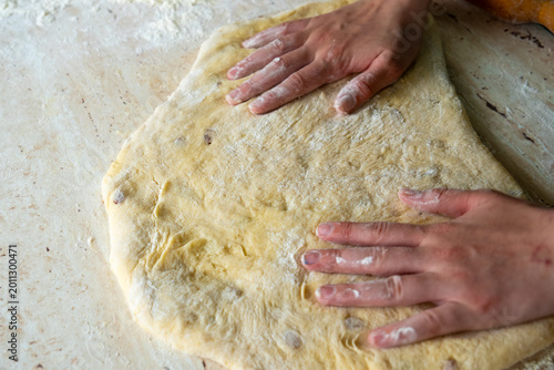 Human palms press and stretch elastic yellow pastry mass containing raisins on flour dusted marble surface for traditional homemade bread preparation