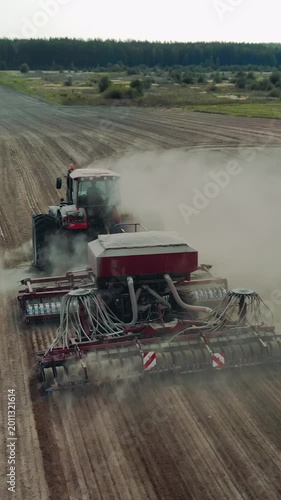 Vertical drone shot of a heavy tractor pulling a modern sowing machine through a dry field. Clouds of dust rising as the agricultural machinery performs spring or autumn planting on a large farm