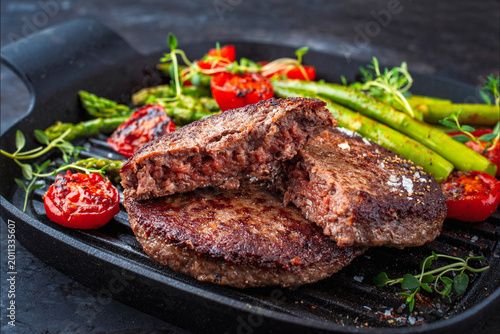Grilled beef patties with roasted green asparagus, tomatos and fresh herbs served as close-up in a rustic design frying pan