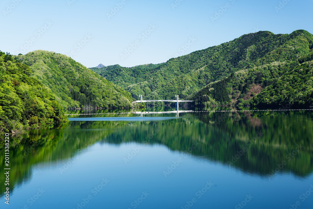 Fototapeta premium Calm Mountain Lake with Fresh Green Forest Reflections and Distant Bridge in Spring