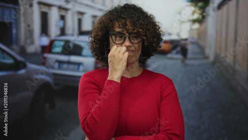 Schilderij op canvas Middle aged hispanic woman touching her forehead with hand, wearing glasses and a red sweater on a narrow cobblestone city street with parked cars; thinking concerned