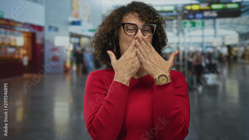 Photos Woman wearing red sweater and glasses covers mouth with both hands at airport terminal; surprise reaction