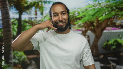 Man with hand to ear for call and pointing finger in forest with green foliage and white t shirt; friendly connection.