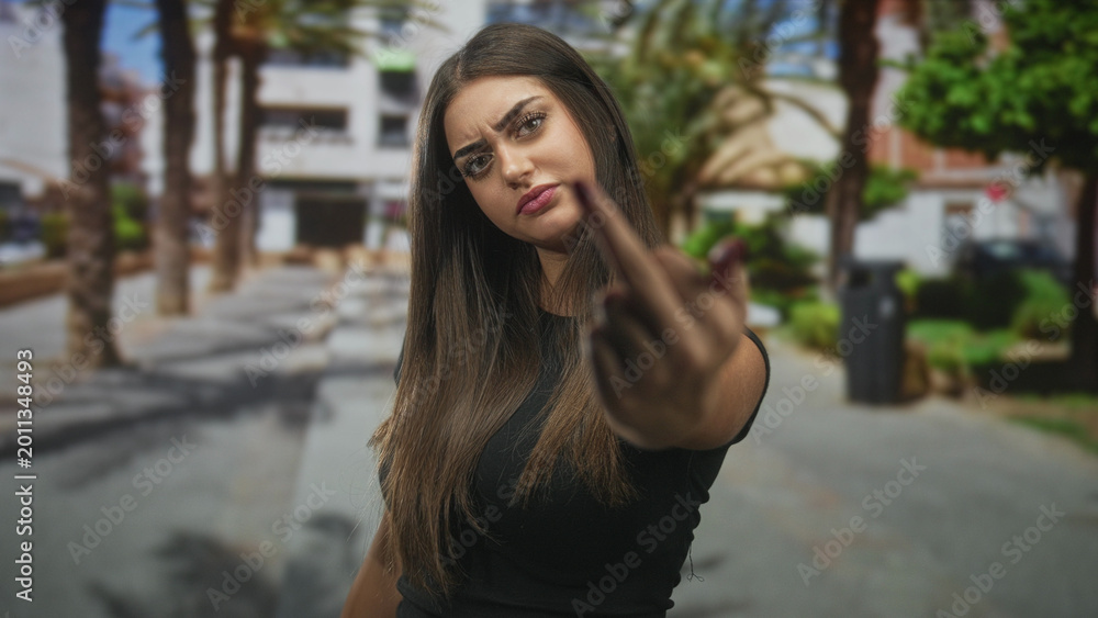 Obraz premium Young hispanic woman showing middle finger and extending hand toward camera on a sunny urban street with palm trees; defiance.
