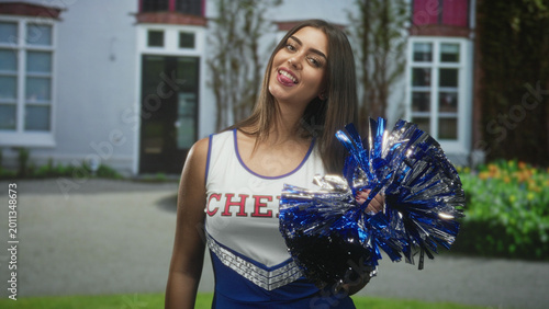 Billede på lærred Cheerleader woman young hispanic smiling and holding a blue metallic pompom with hand at chest in front of a building entrance; team spirit joy