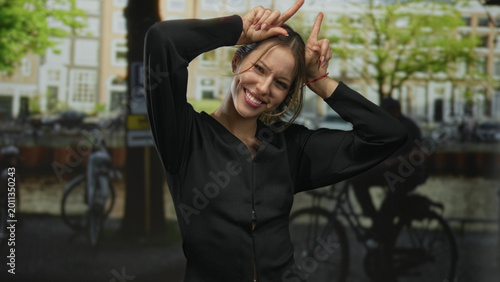 Photos Woman showing horns gesture with hands and smiling face on a street in amsterdam; playful mischief moment