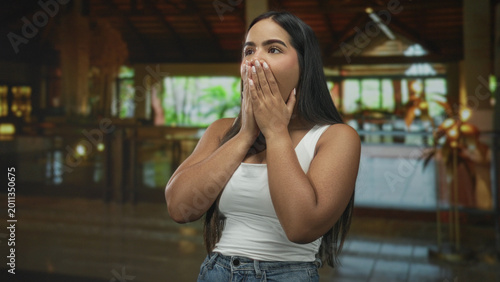 Schilderij op canvas Woman pointing finger with mouth open, wearing white tanktop and denim jeans, standing in a hotel lobby building near reception desk and plants, expressive gesture; surprise