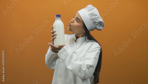 Fotografie Woman chef holds milk bottle with both hands, mouth open in a playful kiss or singing gesture, studio setting with orange backdrop; dairy pride joy