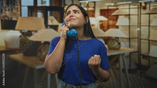 Cuadro en lienzo Woman holding blue handset to ear, eyes closed with fist raised and open hand gesture in a lamp showroom building; melancholy longing